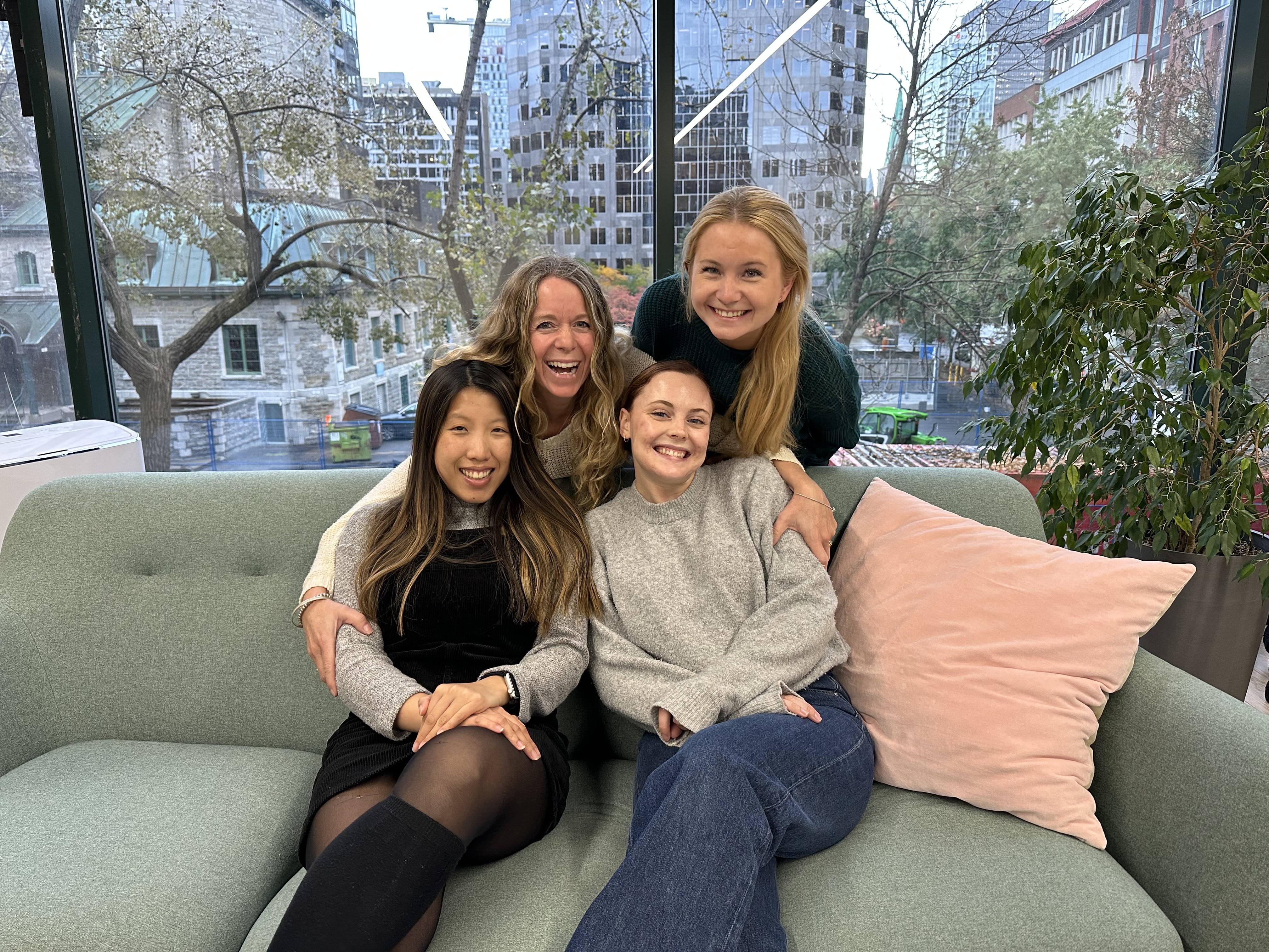 four women smiling with two sitting on a couch and two behind it.